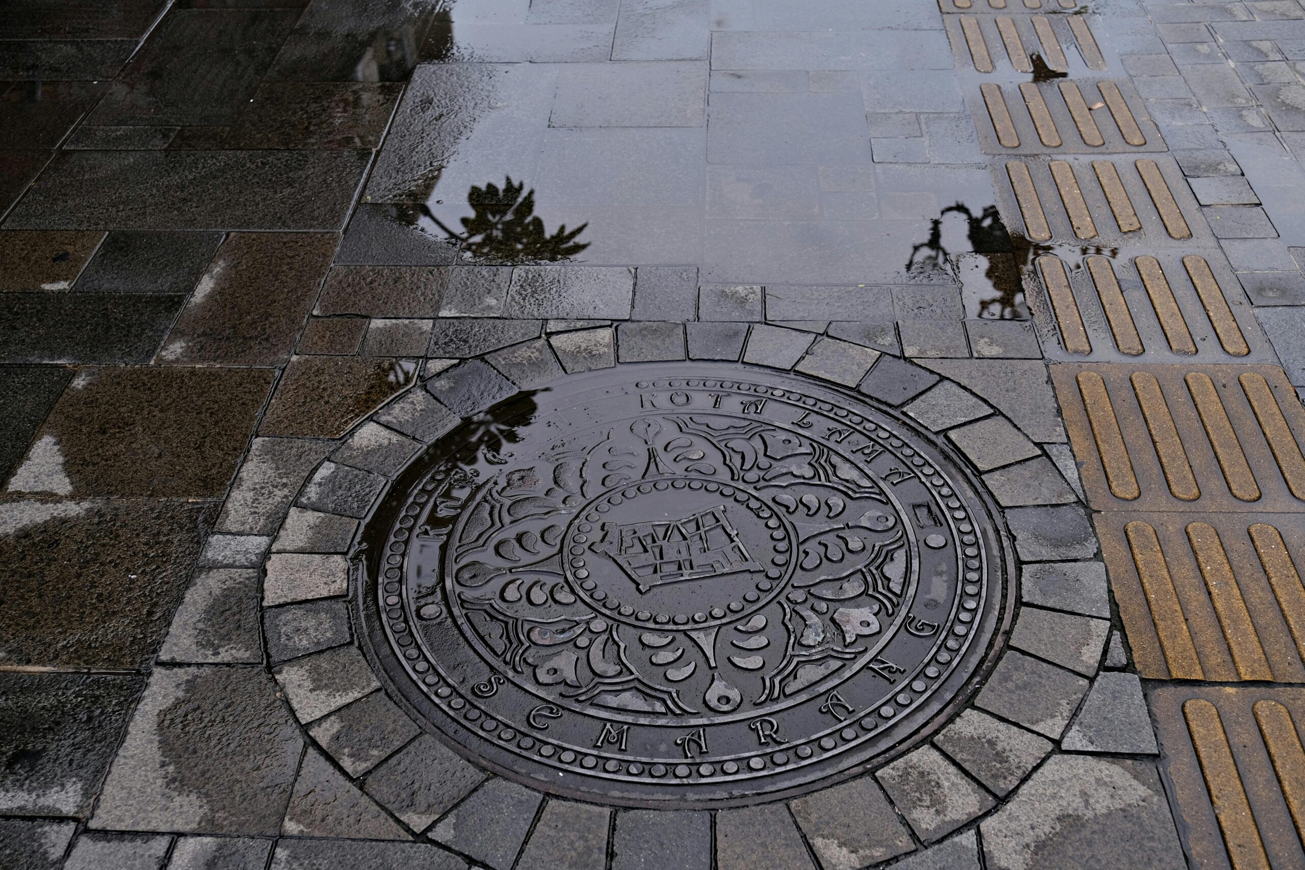 An artistic view of a decorated manhole cover in Semarang, featuring wet pavement reflections.